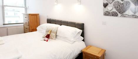 A neatly made bed with white linens, a wooden nightstand with drawers, and a wall-mounted light fixture.