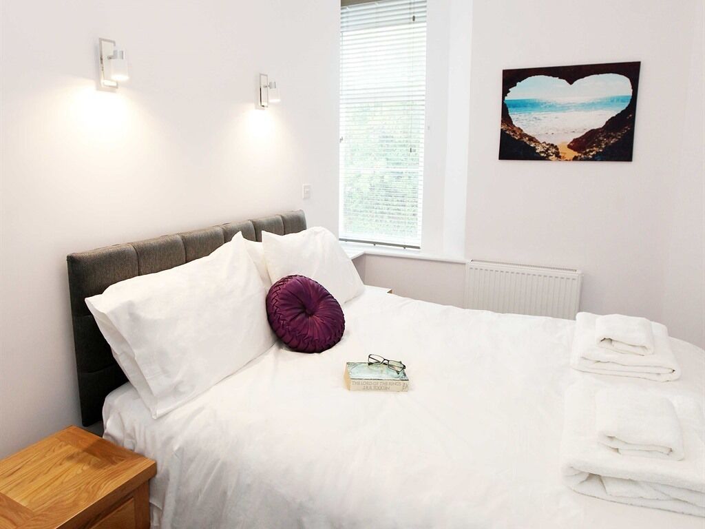 Een net opgemaakt bed met witte lakens, een paars kussen en een boek op het bed. Er is een raam met zonwering, een aan de muur bevestigd fotolijstje en een klein houten nachtkastje naast het bed.