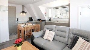 A modern living room with a grey sofa, wooden coffee table, and a small plant. A kitchen with a dining area is visible in the background. - Austen's Luxury Apartments (Torquay)