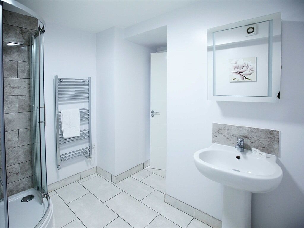A modern bathroom with a glass shower enclosure, a towel rack, a sink with a marble backsplash, and a framed picture on the wall.