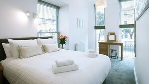 A neatly arranged bedroom with a large bed, a desk, and a chair, along with a window providing natural light. - Austen's Luxury Apartments (Torquay)