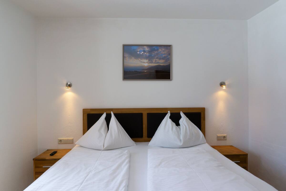 Uma cama de casal com lençóis brancos e cabeceira de madeira, duas mesas de cabeceira, um quadro na parede e duas arandelas.