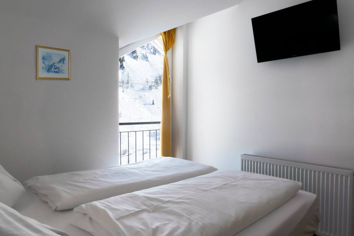 Quarto com duas camas, janela com vista para uma paisagem nevada, quadro na parede e televisão de tela plana presa acima das camas.
