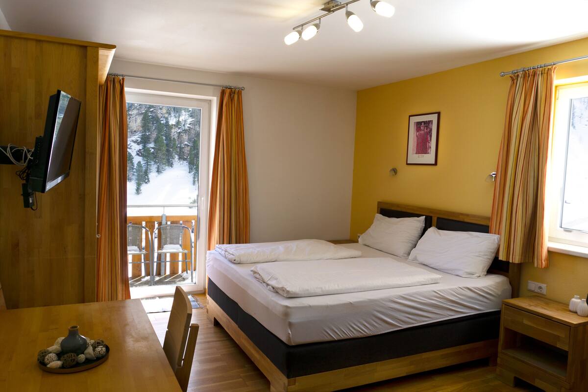 Quarto com cama, televisão na parede, mesa com um vaso e janela com cortinas.