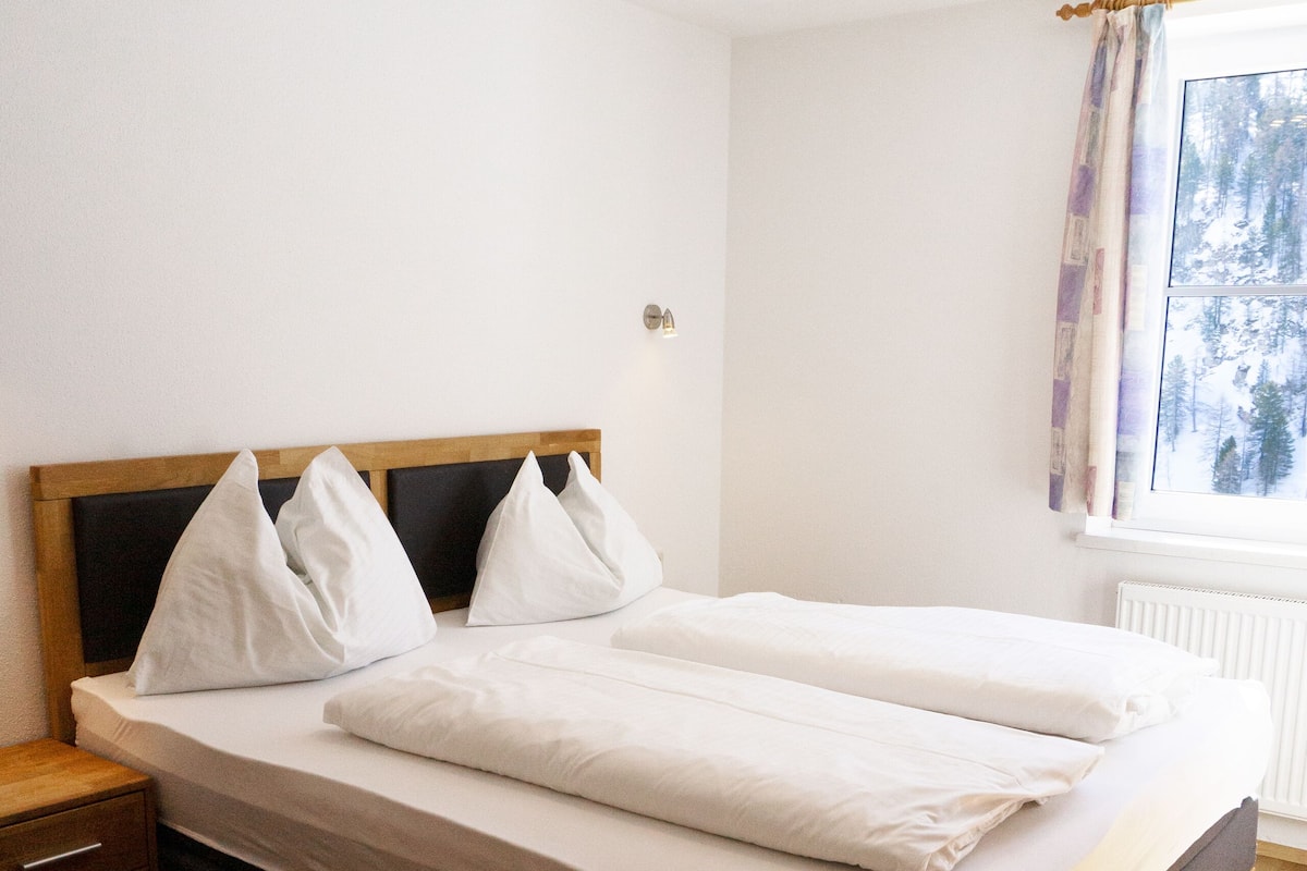 Uma cama de casal com cabeceira de madeira, roupa de cama branca e uma mesa de cabeceira com gavetas.