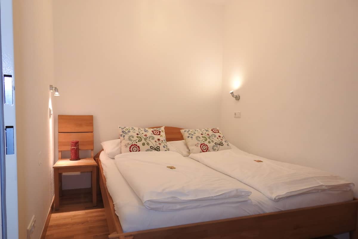 Uma cama de solteiro com cabeceira de madeira, duas almofadas com estampas florais, uma mesa de cabeceira com uma vela vermelha e um luminário fixado na parede.