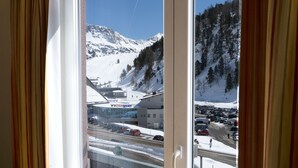 Une fenêtre avec des rideaux, une vue sur une montagne enneigée, un parking et un bâtiment portant l’inscription « INTERSPORT ».