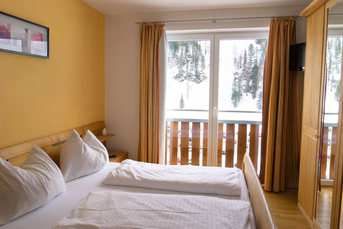 Quarto de hotel com cama, almofadas, cabeceira de madeira, mesa de cabeceira, televisão e varanda com cortinas.