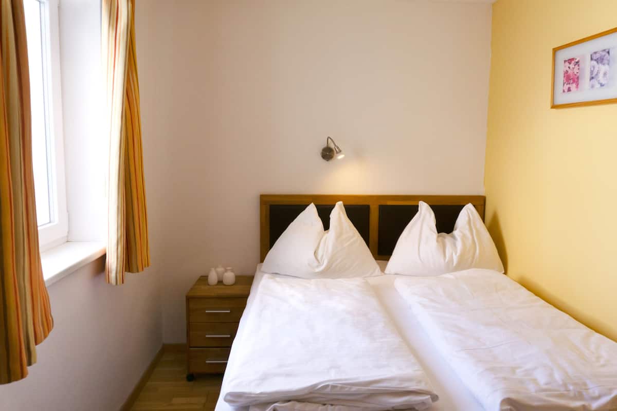 Quarto de hotel com cama, mesa de cabeceira, luminária e janela com cortinas.