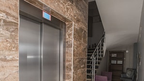 Een hal met een marmeren wand, een lift, een trap en een zithoek met een paarse stoel en een klein tafeltje.
