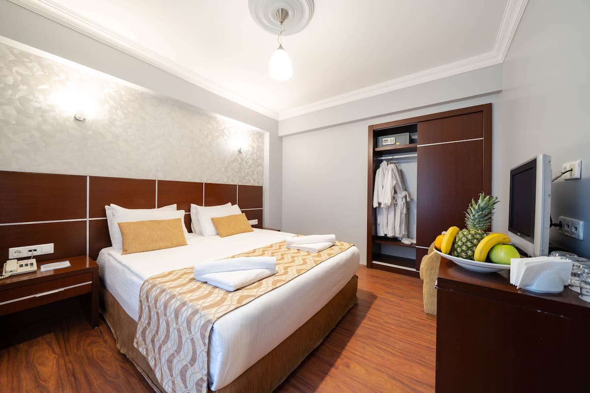 Ein Hotelzimmer mit einem großen Bett, einem hölzernen Kopfteil, einem Nachttisch mit Telefon, einer Obstschale und einem Fernseher auf einem Holzgestell.