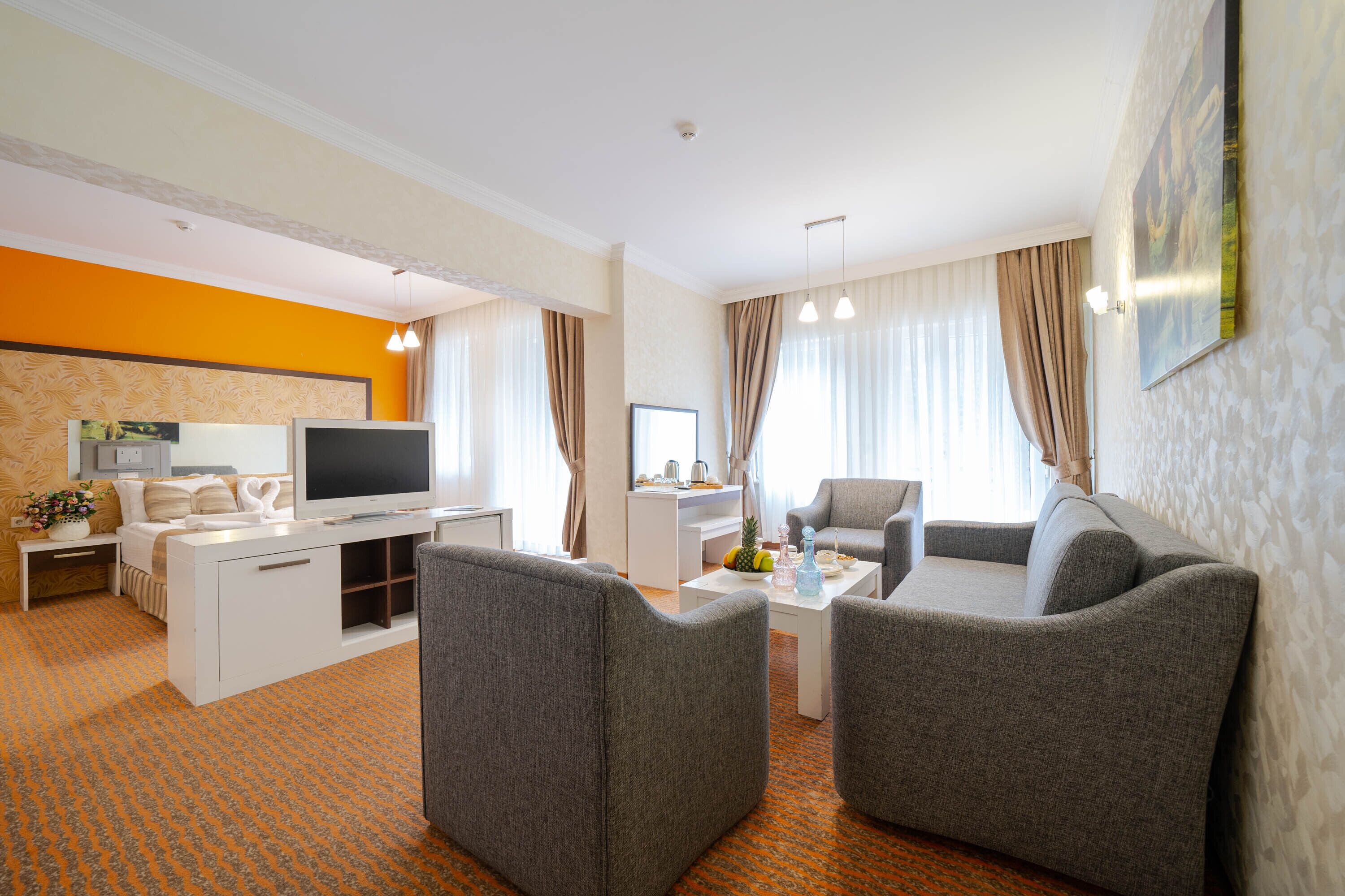Ein modernes Hotelzimmer mit einem Flachbildfernseher, grauen Sesseln, einem weißen Sofa, einem Couchtisch und einem großen Fenster mit Vorhängen.