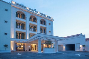A multi-story hotel with a classic architectural design, featuring a central entrance with a covered walkway and a parking area in front.