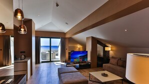 Habitación de hotel moderna con televisor grande, asientos cómodos, balcón con vista al mar y zona de comedor.