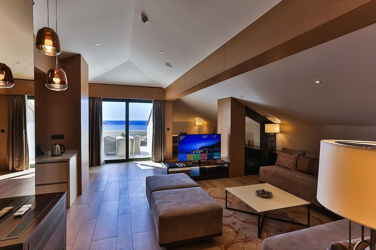 A modern hotel room with a large TV, comfortable seating, a balcony with a view of the sea, and a dining area.
