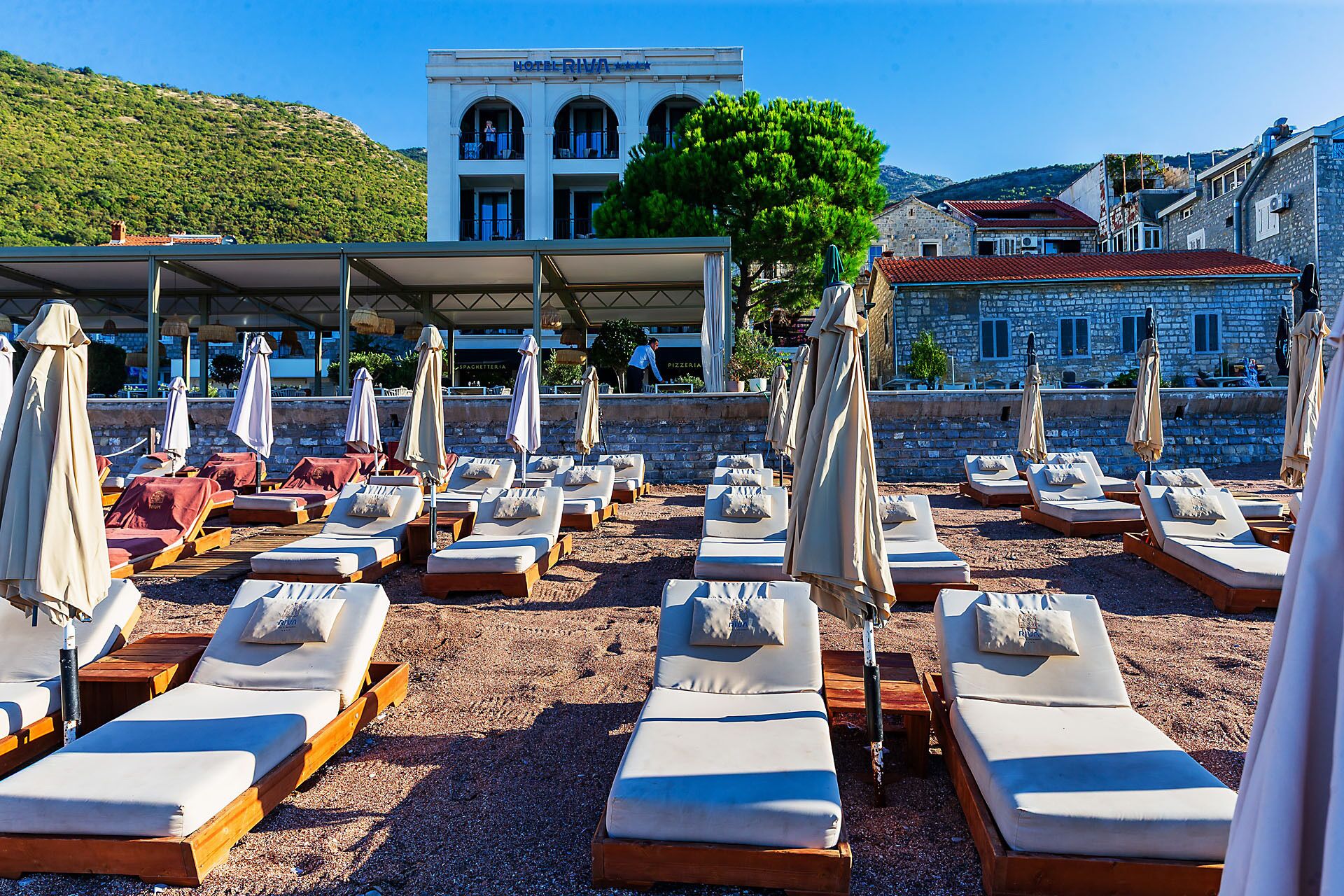 Un hôtel situé en bord de mer, doté de chaises longues et de parasols, un bâtiment en pierre avec des arcades, et une vue sur la mer et les collines.