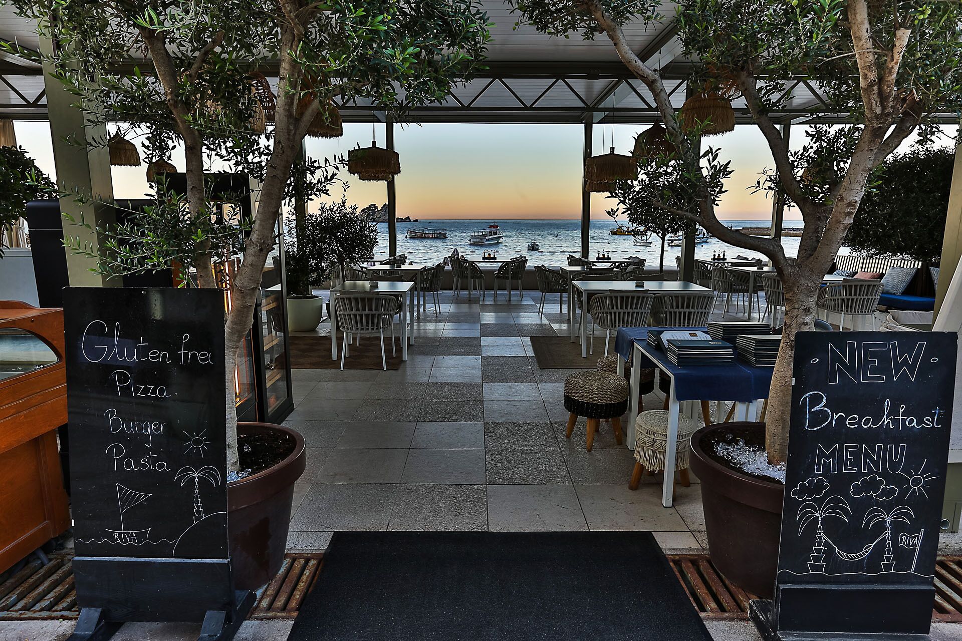 An outdoor dining area with tables and chairs, overlooking the sea. There are signs indicating gluten-free and new breakfast menu options.