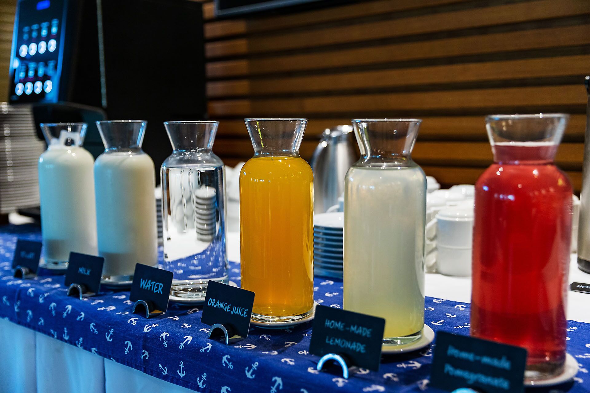 Una estación de bebidas con contenedores etiquetados para leche, agua, zumo de naranja, limonada y zumo de granada.