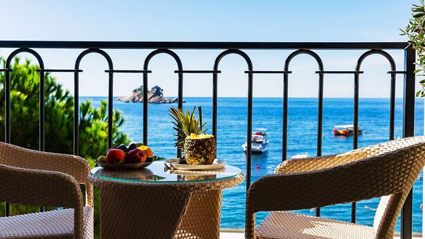 Un balcón con sillas de mimbre, una mesa con fruta y vista al mar con barcos.