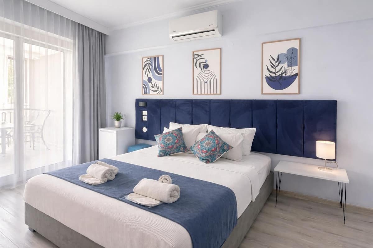 A hotel room with a large bed, a bedside table with a lamp, a wall-mounted air conditioner, and three framed artworks.