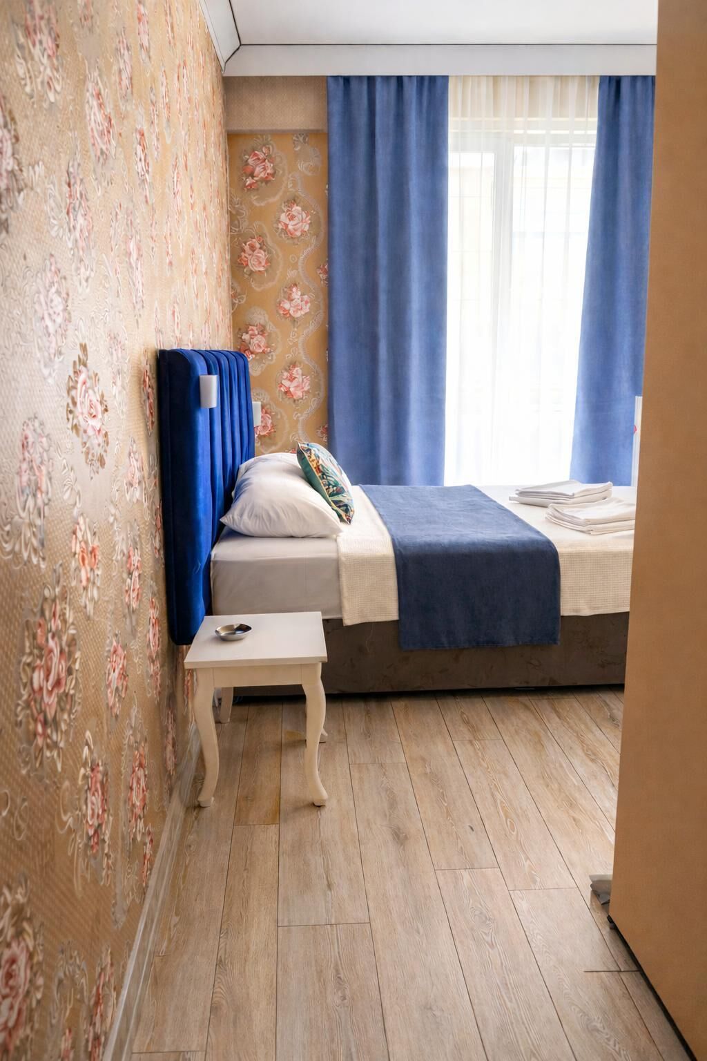 A bedroom with a bed, a small bedside table, a window with curtains, and floral wallpaper.