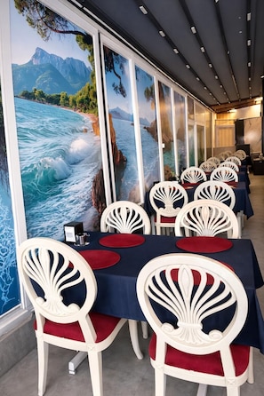 Un coin repas avec des chaises blanches et des coussins rouges, une longue table et une fresque représentant un paysage cÎtier.