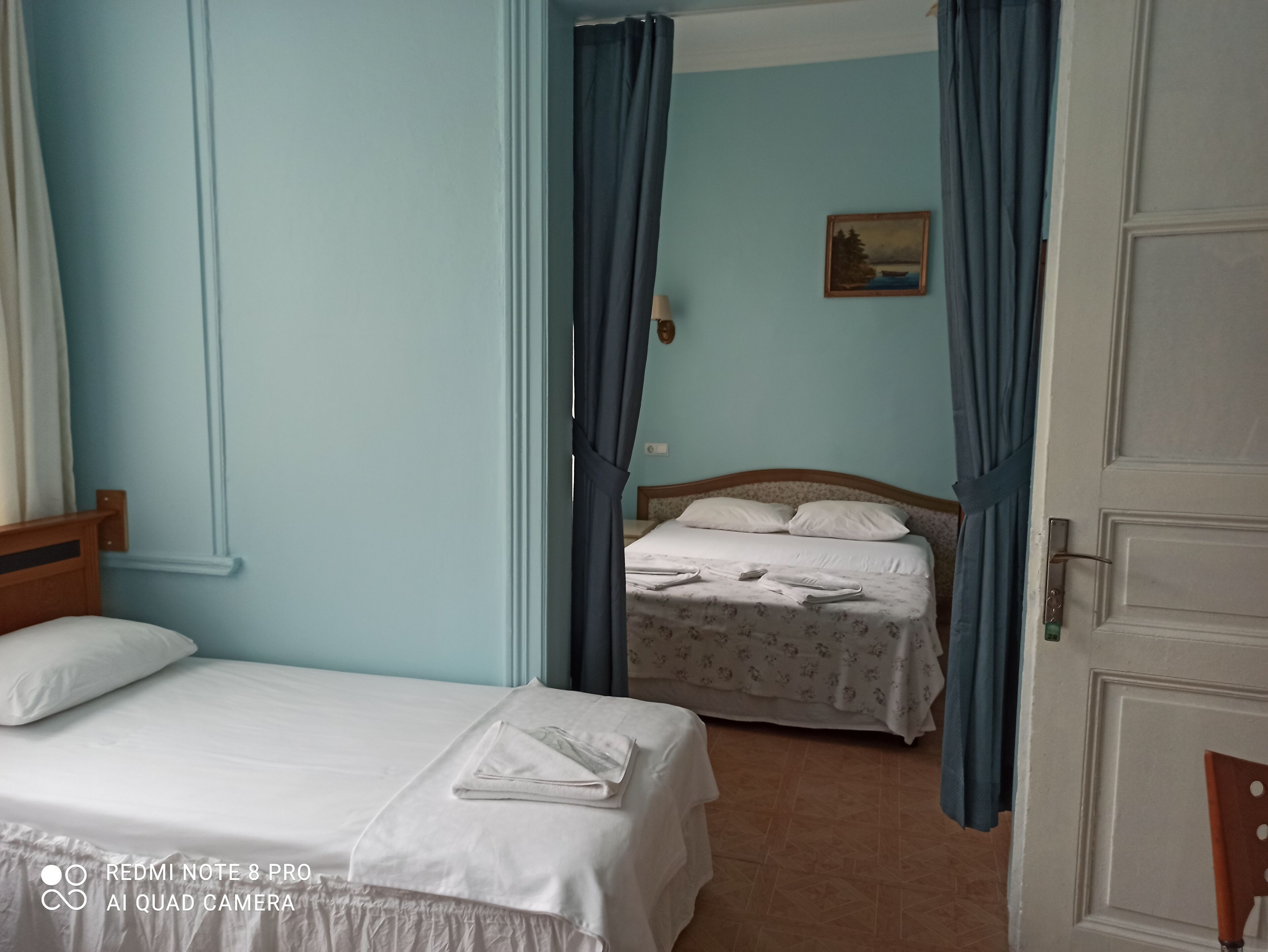 Habitación de hotel con una cama individual, otra cama al fondo y un cuadro en la pared.