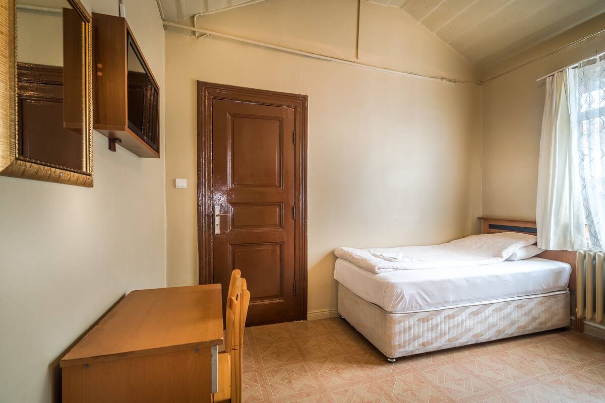 A small, clean room with a wooden desk, a single bed, and a door.
