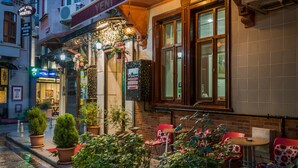 A cozy outdoor seating area with potted plants and red chairs, a visible entrance to a building with a sign reading "HOTEL," and a window with a view of the street. - Yeni Hotel (Istanbul)