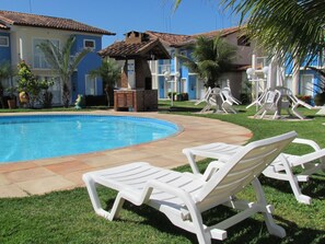 Uma piscina com espreguiçadeiras, um gazebo de tijolos e um gramado bem cuidado.