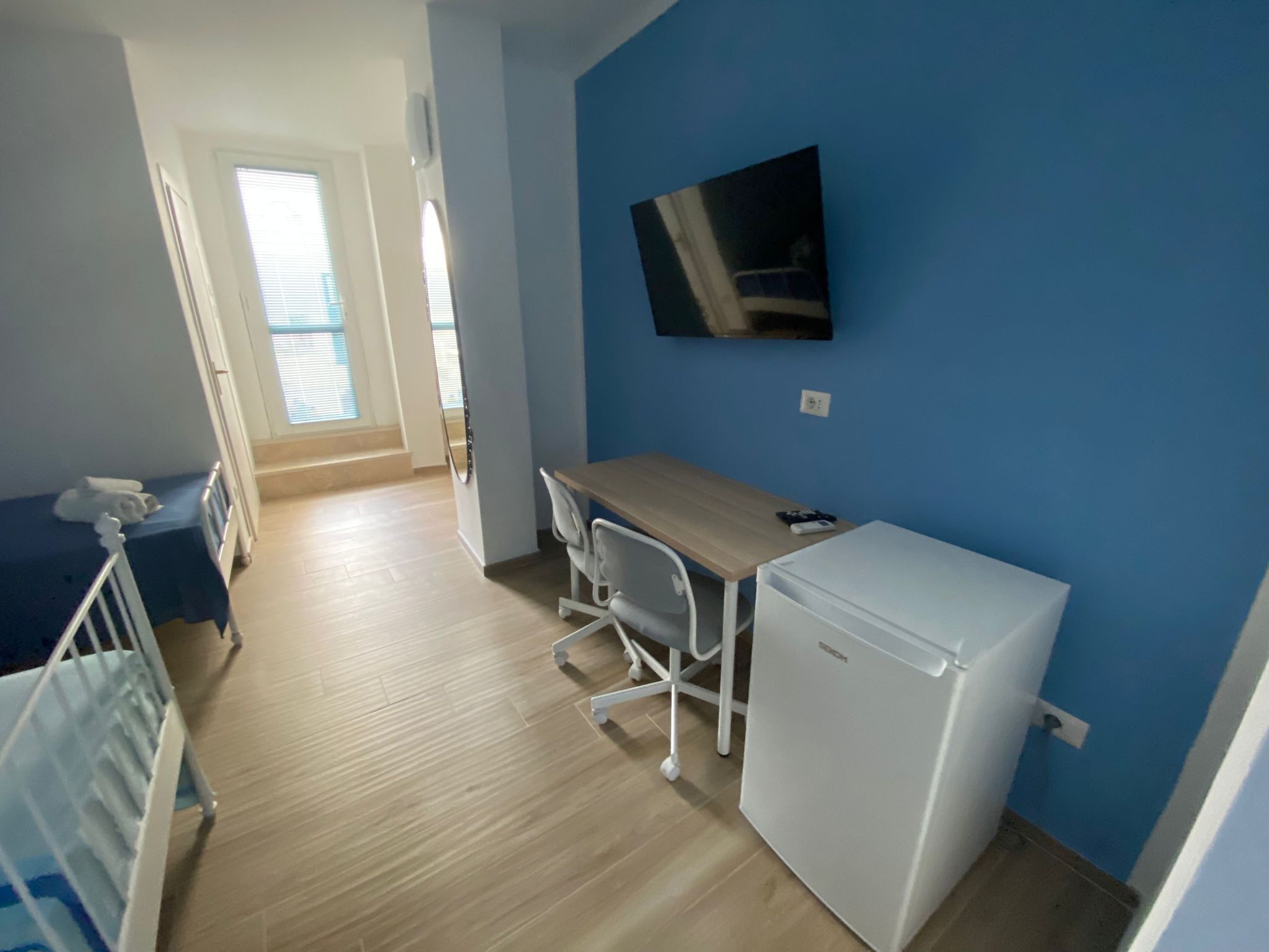 A compact room with a wooden floor, a small desk with a chair, a white refrigerator, and a TV mounted on the wall.