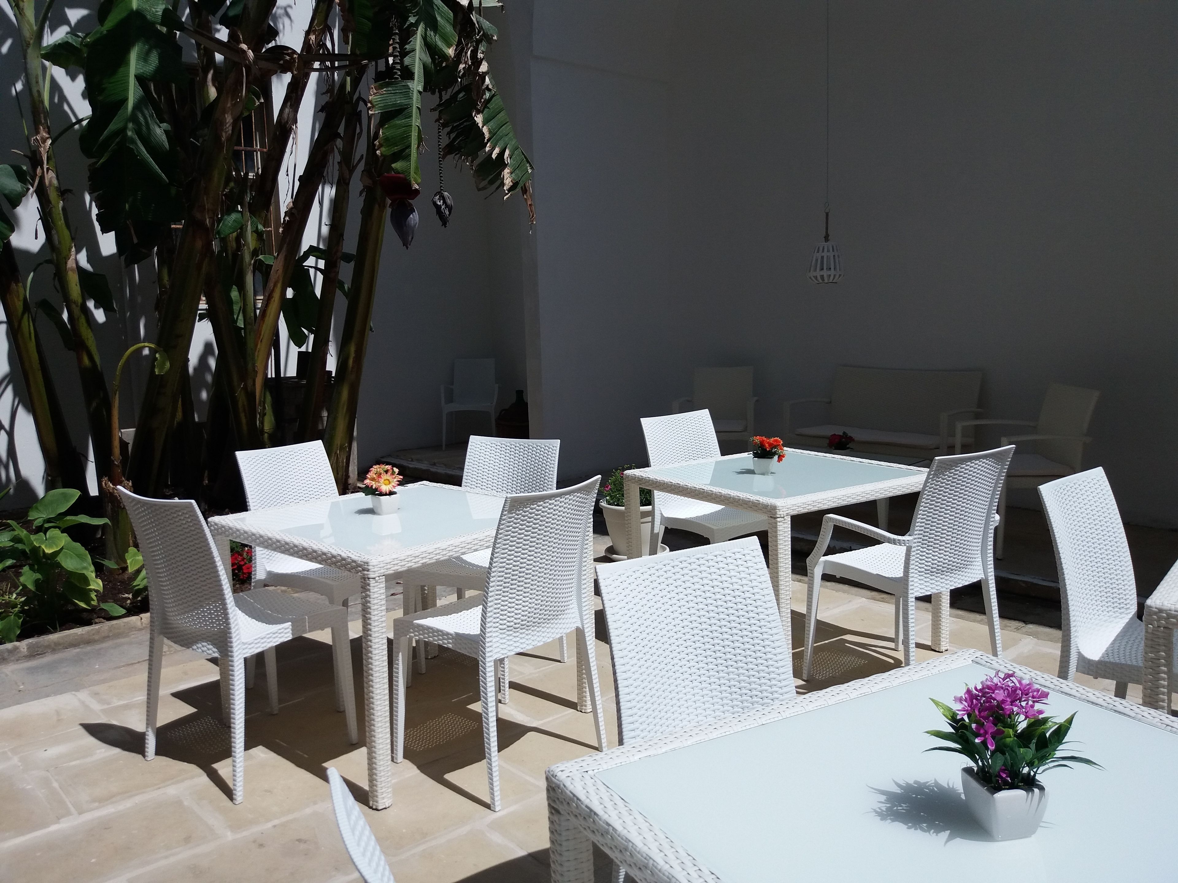 An outdoor patio with white wicker furniture, including tables and chairs, surrounded by potted plants and a large tree.