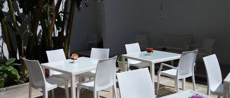 An outdoor patio with white wicker furniture, including tables and chairs, surrounded by potted plants and a large tree.