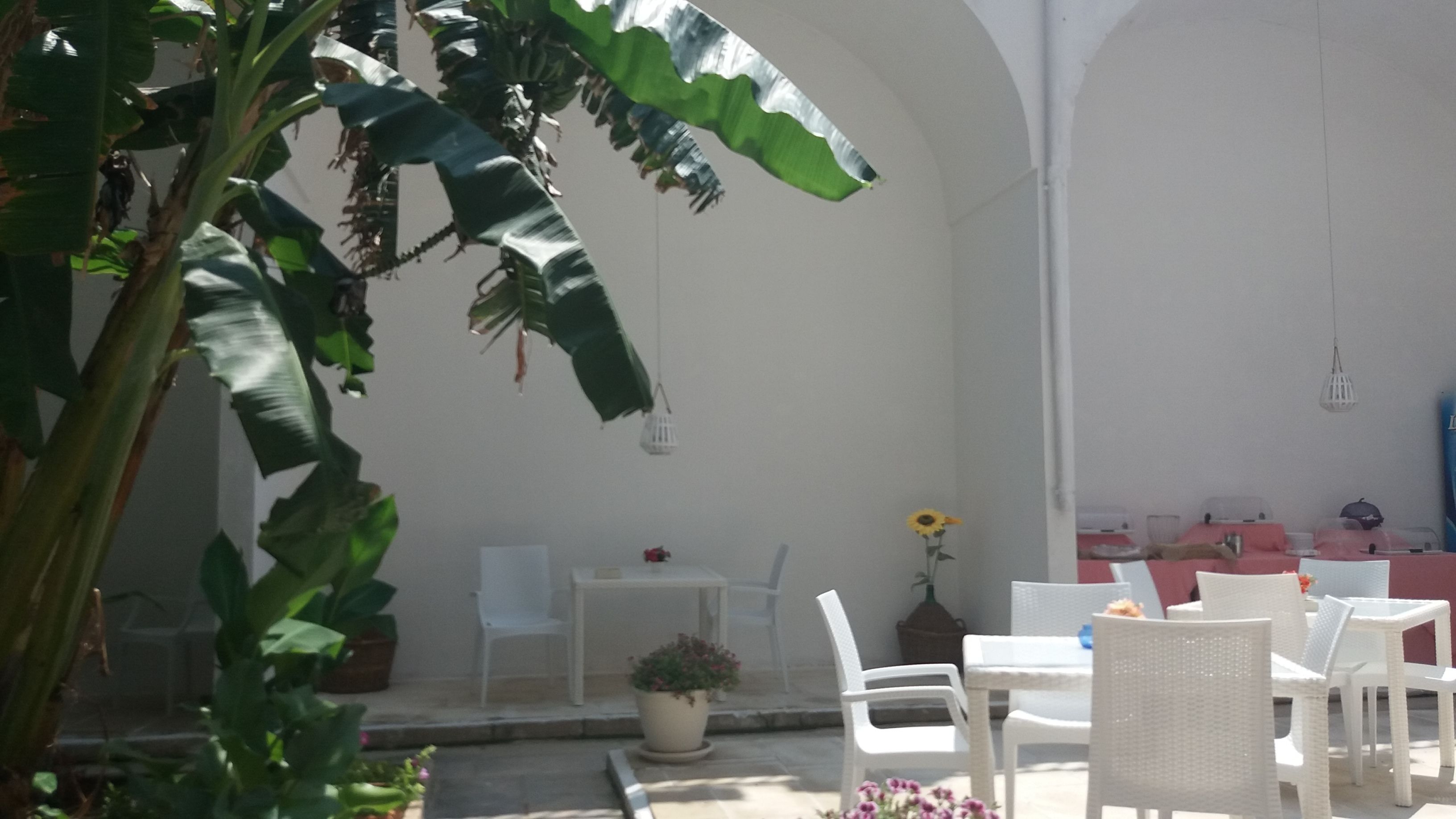 Un patio al aire libre con mobiliario de mimbre blanco, una pequeña mesa con una planta en maceta y una pared iluminada por el sol.