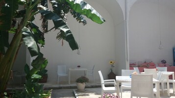 Un patio al aire libre con mobiliario de mimbre blanco, una pequeña mesa con una planta en maceta y una pared iluminada por el sol.