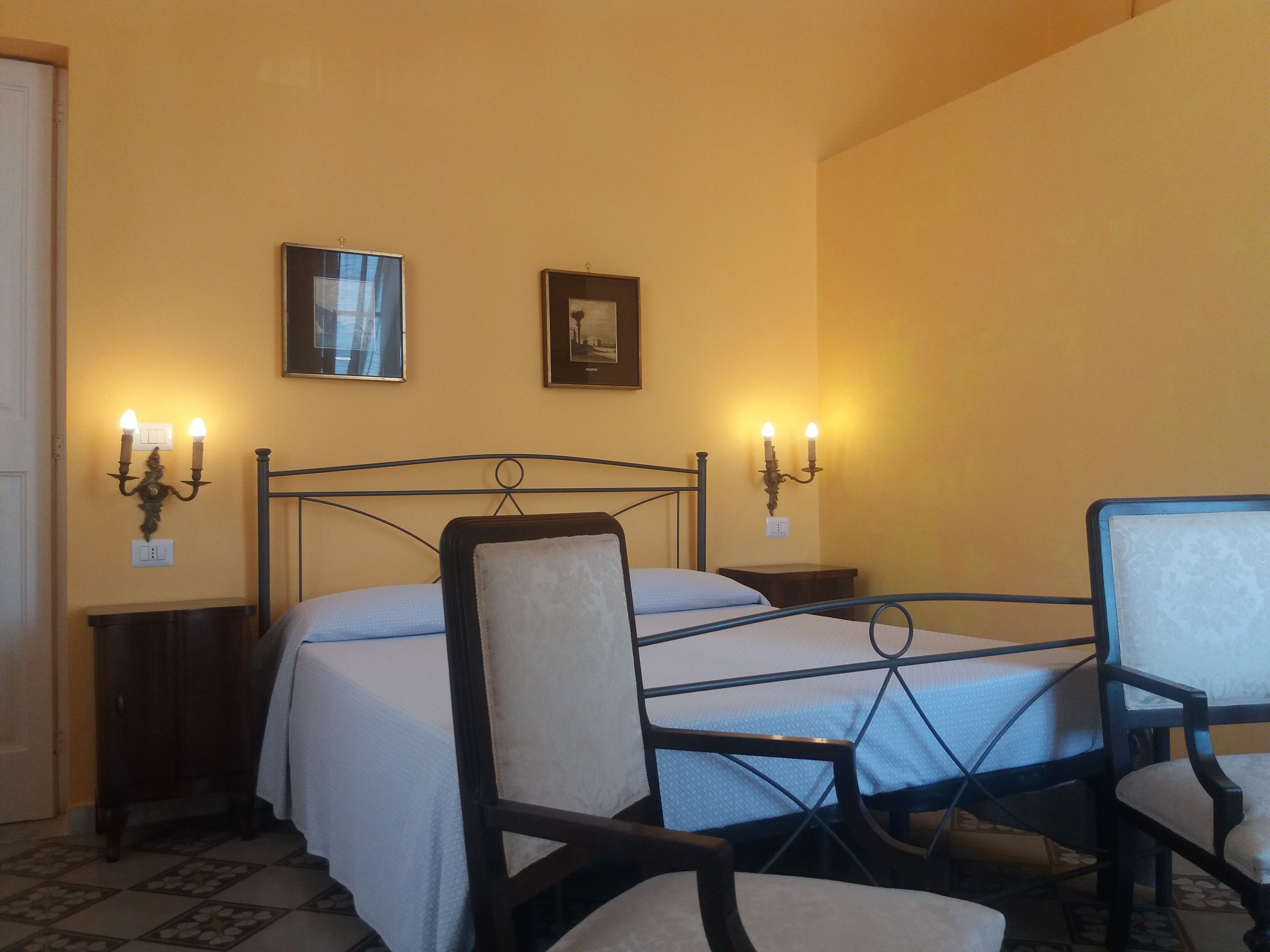 A bedroom with a bed, two wall-mounted sconces, a chair, and two framed pictures.
