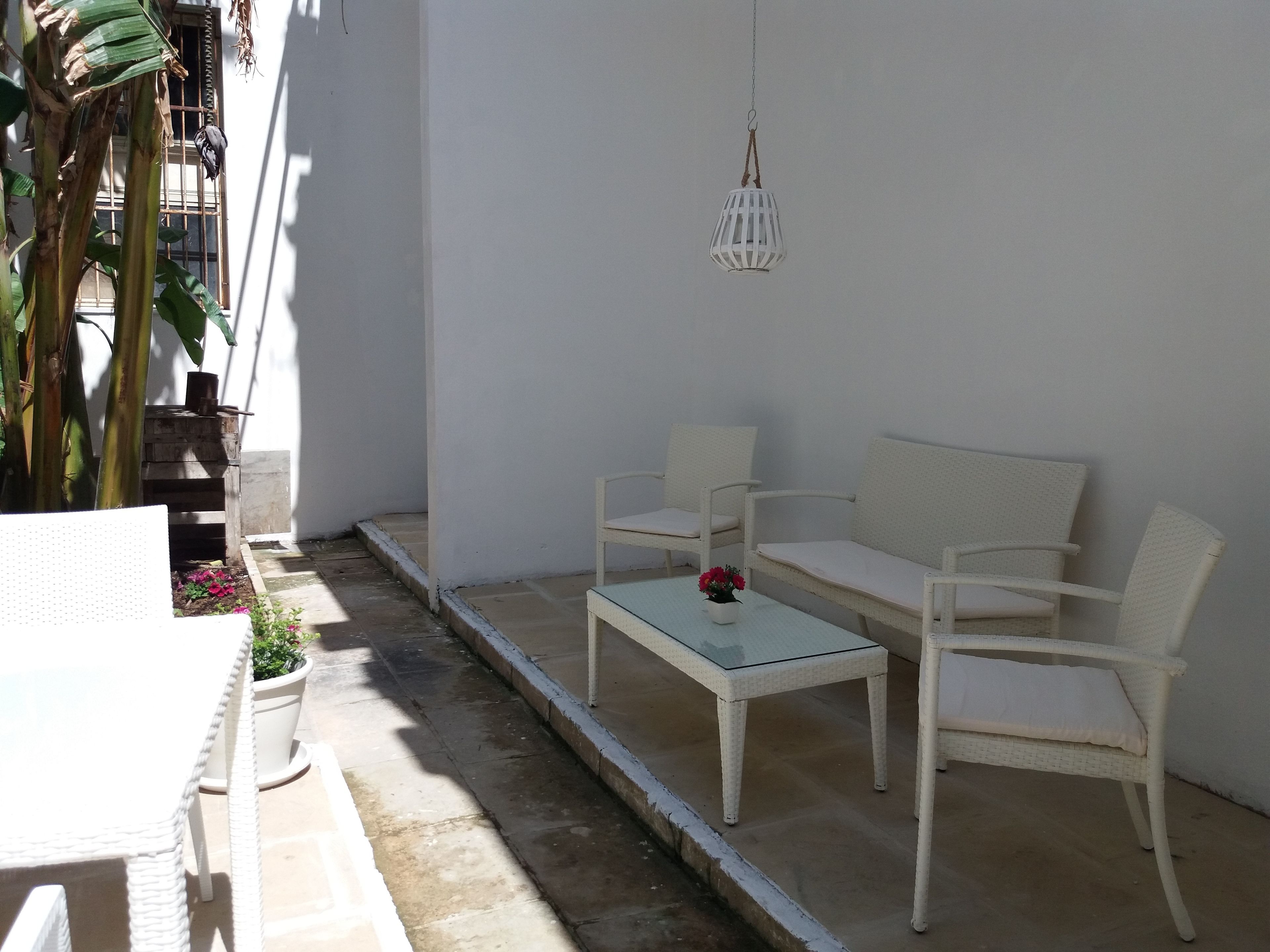 Un espacio exterior con mobiliario de mimbre blanco, una mesa de vidrio, una lámpara colgante y una planta en maceta.