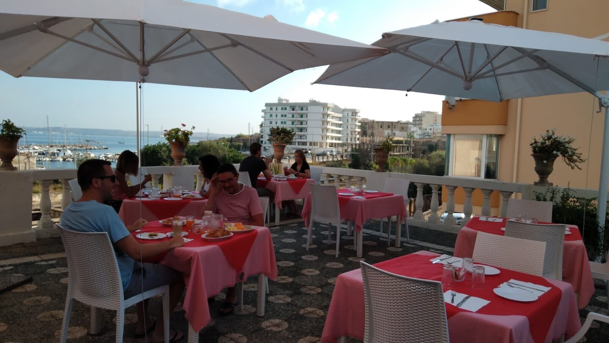 Un área de comedor al aire libre con mesas, sillas, sombrillas y vistas al mar y edificios al fondo.