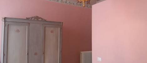 Une pièce aux murs roses, avec un radiateur, une chaise blanche et une armoire ornée de motifs floraux.