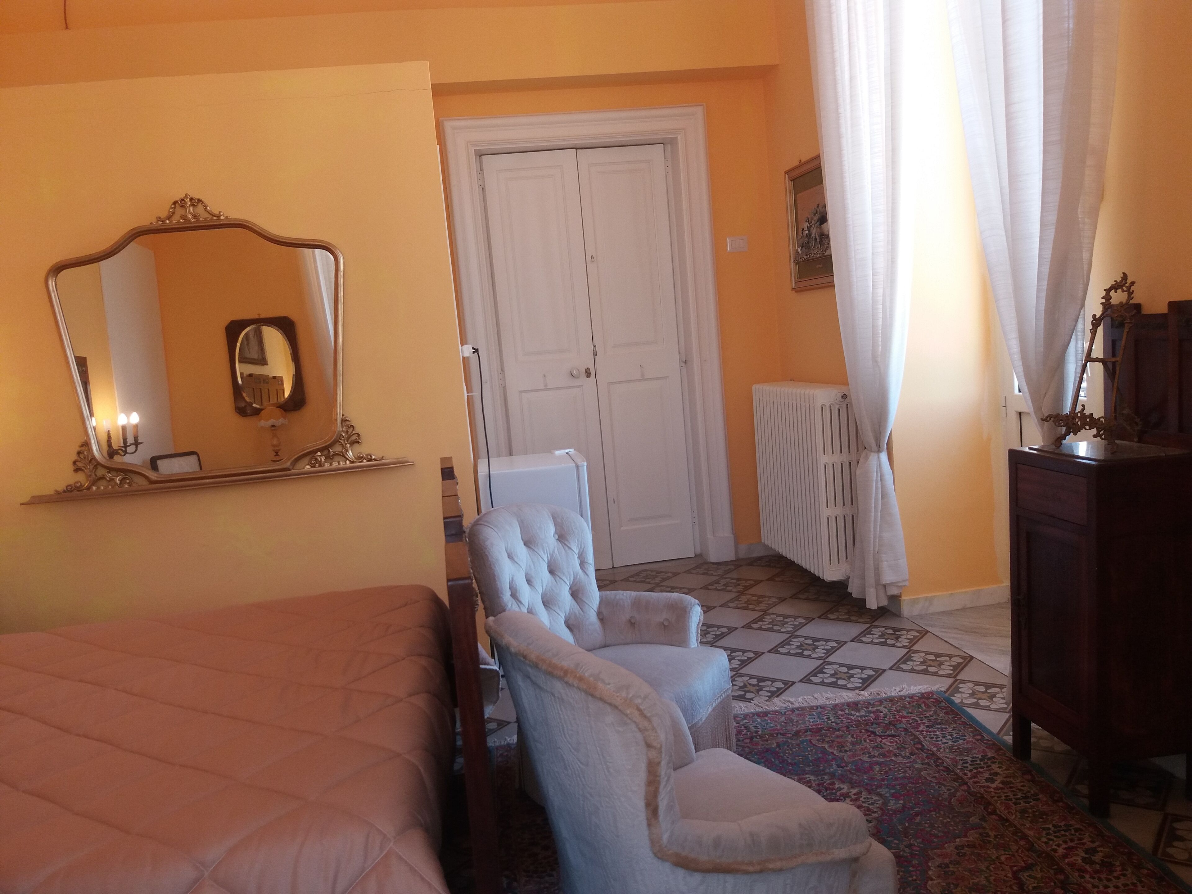 A bedroom with a bed, two armchairs, a dresser, a mirror, a radiator, and a door.