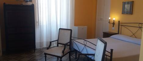 A bedroom with a bed, two chairs, a desk, a dresser, and a window with curtains.