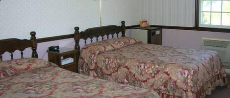 Una habitación con dos camas de madera, ropa de cama floral, una mesita de noche con lámpara y una ventana con persianas.