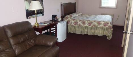 A bedroom with a brown leather sofa, a small desk with a lamp, a bed with floral bedding, and a window with a valance.