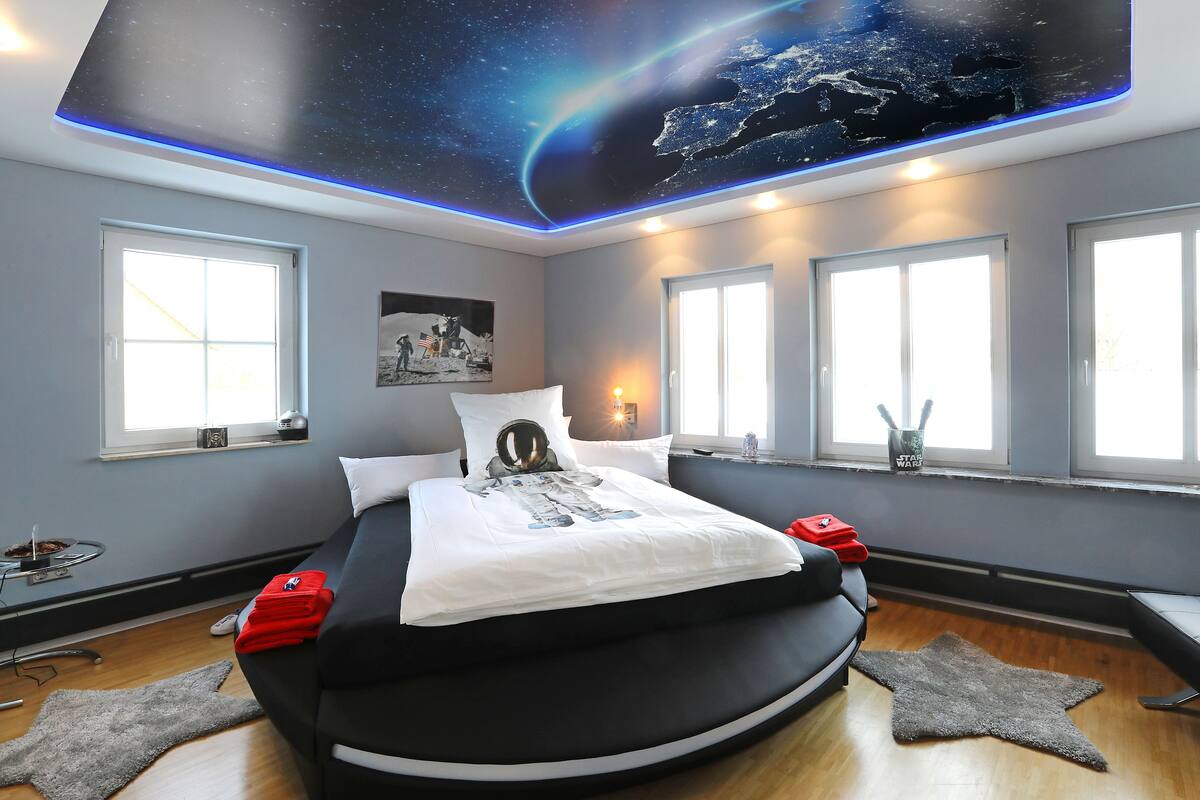 Ein Schlafzimmer mit Bett, einer Decke im Weltraumdesign, großen Fenstern und einer Star Wars-Tasse auf der Fensterbank.