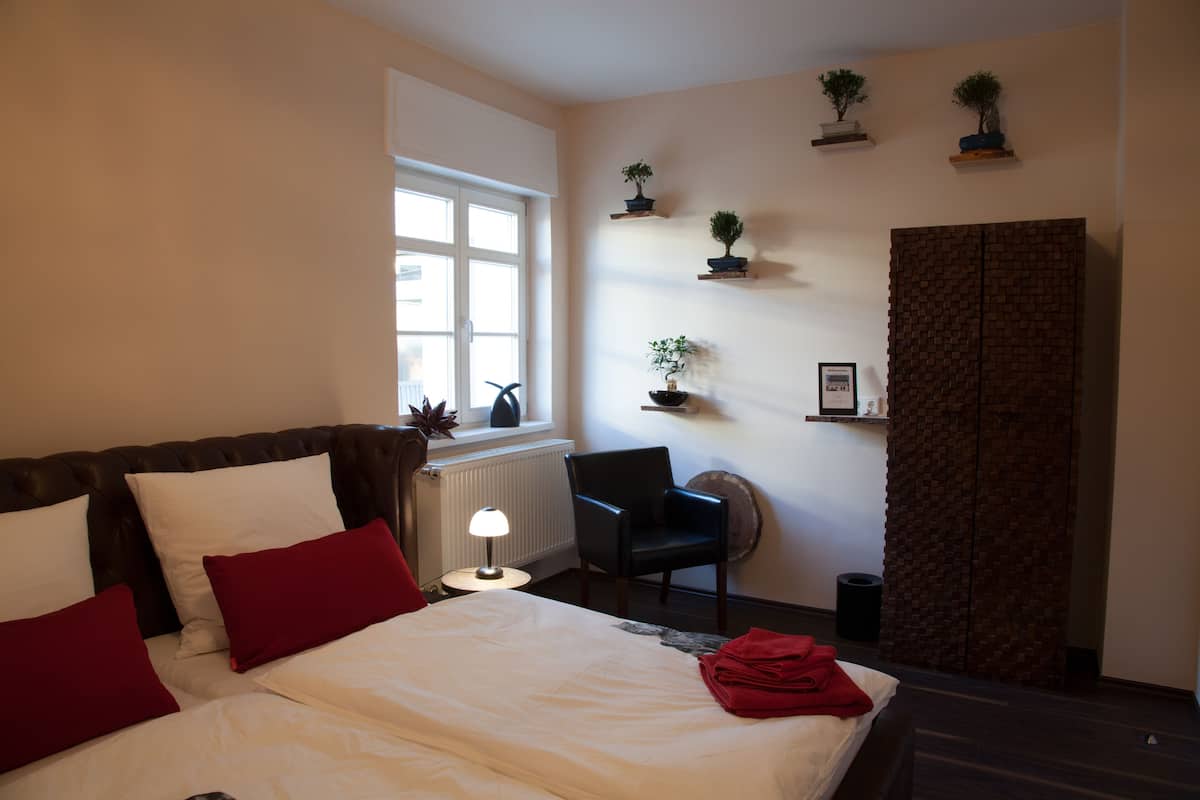 Ein Hotelzimmer mit Bett, Sessel, Heizkörper, Fenster und Wandschränken mit Blumentöpfen.