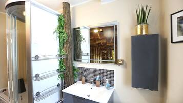 Un baño con cabina de ducha de vidrio, un lavabo blanco con salpicadero de azulejos, un espejo y una planta en maceta.