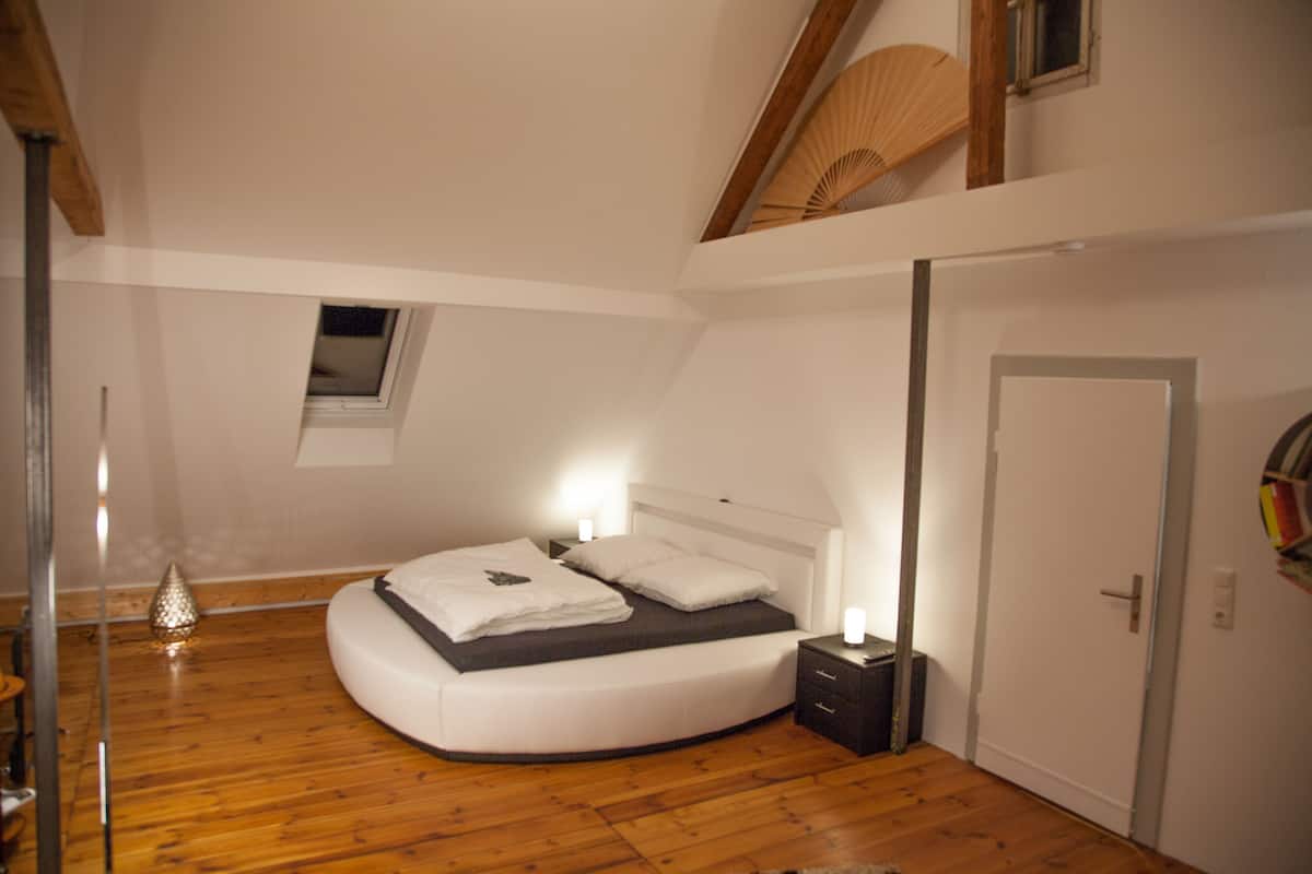Ein Schlafzimmer mit einem gebogenen Bett, Nachttischlampen, einem kleinen Fenster und Holzboden.