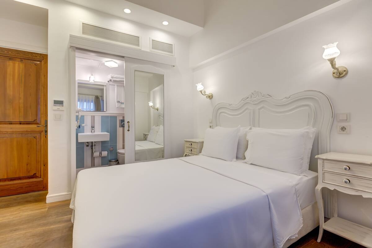 A bedroom with a large bed, a wooden door, a bathroom with a sink and mirror, and wall-mounted lights.