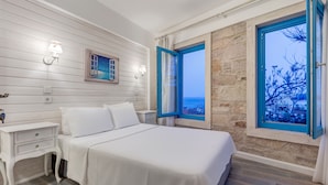 Un dormitorio con una cama grande, mesitas de noche blancas, una ventana con contraventanas azules y una pared de piedra.