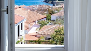 Vista desde una ventana hacia un pueblo costero con edificios de tejados de terracota, una fortaleza histórica y el mar.